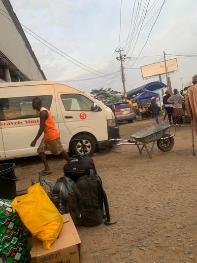In Elelenwo, Nigeria, a street scene unfolds under a cloudy sky, with a white van labeled "Travels Shut" parked on the left. A Black man in an orange tank top and brown shorts walks from left to right beside the van. In the foreground, several pieces of luggage are visible, including a yellow plastic bag, a cardboard box, and two backpacks. A wheelbarrow, marked with "GOD IS," is positioned in the middle of the dirt and gravel road. Further in the background, several people are gathered near stalls and another vehicle. Overhead, a network of electrical wires crisscrosses the sky, and a large, blank billboard stands on a pole. The environment appears to be a bustling outdoor market or transport hub.