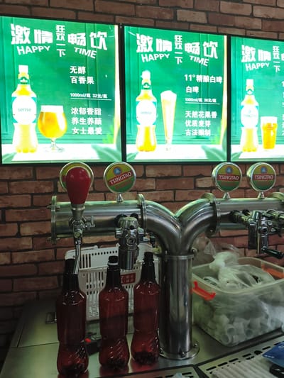 The image shows a beer dispenser with multiple taps in what appears to be a bar or restaurant. Three illuminated signs are visible behind the dispenser, each displaying Chinese characters and images of bottles and glasses of beer, suggesting different beer offerings. The signs are predominantly green and white, with some red accents. The beer dispenser itself is made of polished metal and has several taps, each with a branded badge, including "TSINGTAO". Below the dispenser, several empty brown plastic bottles are lined up, ready for filling. In the background, a brick wall is visible. There are no people in the image. The location is identified as Jixi, China.