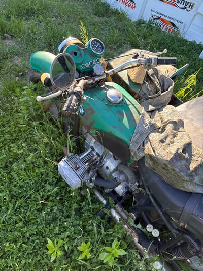 The image shows a partially disassembled green motorcycle lying on its side in a grassy area. A large rock and fabric scraps are resting on the motorcycle's fuel tank and seat. The motorcycle's engine is visible, as are gauges, handlebars, and a mirror. The immediate environment is characterized by green grass and weeds. In the background, there is a white box with the words "HAUSTHERM" printed on it. The location of the image is not discernible from the image's visual contents.