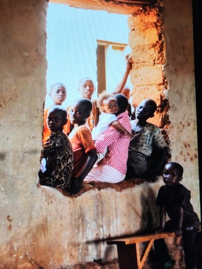 The image displays eight individuals, predominantly children and young adolescents, situated within a building in Dodoma, Tanzania, featuring rough, unplastered earthen walls. Six individuals are positioned within a large, unglazed opening, illuminated by bright natural light from an exterior source. From left to right within this opening: a child wearing a leopard-print long-sleeved top; a child in an orange short-sleeved top; a young person in a red and white checkered shirt holding an infant with light, curly hair and a white top; a young person in a dark, patterned short-sleeved top; and two additional young individuals in orange tops partially visible behind the front row. The majority of individuals in this group, including the infant, are looking towards the viewer; the young person in the dark patterned shirt looks towards the right. An arm, likely belonging to another individual, rests on the earthen doorpost on the right side of the opening. In the lower right foreground, a younger child in a dark, patterned short-sleeved top and dark trousers stands to the left of a small, light-colored wooden bench, looking towards the viewer. The floor surface appears to be earthen.