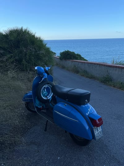 A blue vintage Vespa scooter is parked on a paved road next to a grassy embankment. The scooter is positioned at an angle, facing away from the viewer and slightly to the left. It has a spare tire mounted on the front. In the background, a body of water, likely the sea, stretches to the horizon under a clear blue sky. A low, weathered wall runs along the edge of the road separating it from the embankment and the sea. The scene appears to be in the town of Trabia, Italy, a coastal municipality in the Metropolitan City of Palermo, Sicily. The overall mood is serene and peaceful, suggesting a quiet moment at dusk or dawn.