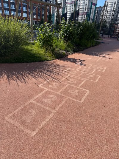 The image shows a children's hopscotch court painted on a reddish-brown, rubberized surface. The court is in the foreground and extends diagonally across the frame. Several squares are visible, with numbers painted in white within them: 1, 2, 3, 4, and 5 are clearly discernible. Beyond the hopscotch court, there is a grassy area with some bushes and trees. In the background, there are modern multi-story apartment buildings and a black metal fence. The scene appears to be an outdoor public space in a residential area. The lighting suggests it is daytime, with shadows cast by the foliage on the hopscotch court. No people are present in the image. The location is identified as Patrusheva, Russia, likely a playground or recreational area within a city.