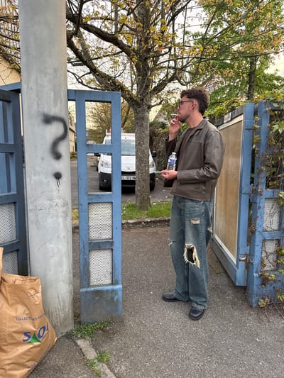 The image captures a man in Orsay, France, standing in front of a blue and grey concrete fence or barrier, smoking a cigarette. The man is wearing a brown leather jacket, ripped blue jeans, and black shoes, and he holds a plastic water bottle in his hand. The environment includes trees with sparse foliage, part of a white van visible behind the barrier with the license plate "CL-372-HW", and a brown paper bag with printed text and a logo positioned near the base of the barrier, next to a section of the fence. Graffiti in the form of a question mark is visible on the concrete portion of the barrier.
