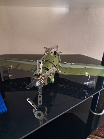 The image captures a close-up view of a detailed model, likely constructed from a Meccano or Erector set, set indoors in Gavar, Armenia. The central subject is an intricate mechanical assembly, predominantly featuring olive-green metal strips and plates connected by numerous screws and nuts. These components are perforated with small holes, characteristic of such construction toys.
The model showcases a central olive-green cylindrical or box-like structure from which broad, flat olive-green blades or arms extend, one prominently on the left. These blades are also perforated and secured with fasteners. In the mid-section of the model, a more complex silver-colored mechanism is visible, comprising several small metal strips, a central shaft, nuts, and what appears to be a coiled spring or stacked washers, suggesting a rotational or articulated joint. Further to the right, another smaller assembly of silver metal strips and a central dark knob or wheel is seen against a dark background.
The setting appears to be an indoor space. A light-colored, textured wall occupies the left side and top-left background. To the right, a dark, glossy, or reflective surface, possibly a piece of furniture made of plastic or dark wood, serves as a backdrop, reflecting some ambient light and showing traces of dust. A curved silver element, possibly a handle or decorative trim, is visible at the top edge of this dark object. A small section of a blue, possibly woven fabric or plastic item, is partially visible at the bottom right corner of the frame. There are no people visible, and no discernible activity is taking place other than the static display of the model. No text is visible in the image. The time of day is indeterminate.