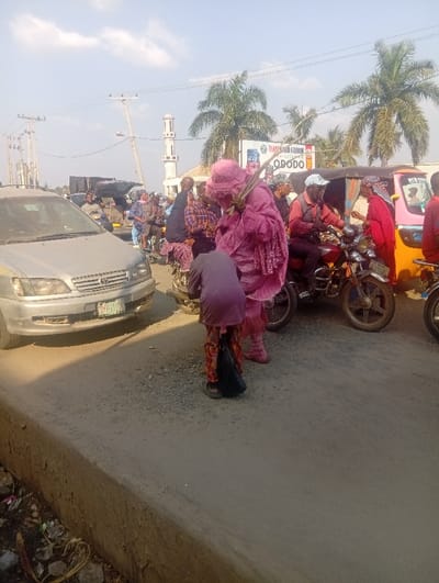 This image captures a busy daytime street scene at Itakpe Junction, Nigeria. A concrete curb occupies the lower foreground, bordering an unpaved, dusty road. On the left, a silver Toyota Sienna minivan is stopped, displaying a Nigerian license plate.

At the center, a figure wears a voluminous, textured pink-purple costume, including a matching head covering that obscures its face. This figure holds multiple thin, stick-like objects vertically in its right hand. Directly in front of this figure, a smaller individual, dressed in a dark purple top and patterned trousers, bends forward, holding a black bag in their left hand.

To the right, a man in a white cap, dark vest, and red shirt is seated on a motorcycle. Further right, a yellow, pink, and green motorized tricycle (Keke Napep) is present, surrounded by several individuals. Numerous other pedestrians and motorcycles are visible throughout the background, indicating active street traffic.

The background features tall palm trees and a white mosque minaret. A large billboard displaying partial text "TEAM AKIN FARIN..." and "ODODO" is visible behind the vehicles. Overhead, utility poles with power lines cross a bright blue sky with scattered white clouds.