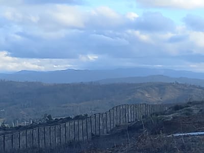 The image presents a panoramic, elevated view of a rural, mountainous landscape in Santa Juana, Chile, under a sky with scattered white and grey clouds against a light blue background. In the immediate foreground, a dark, low-lying slope covered with sparse, dry-looking vegetation is visible. A prominent, dark-colored barrier or fence, constructed from numerous vertical slats or posts, traverses the mid-ground diagonally from the lower-left to the mid-right. This structure appears to follow the contour of a gentle incline. Beyond this barrier, the terrain consists of multiple layers of rolling hills and valleys, covered primarily in muted green, brown, and grey vegetation, indicating a mix of forested areas and possibly cleared or recently disturbed land. The distant background reveals a series of mountain ranges appearing as hazy, bluish silhouettes under the atmospheric perspective. No human figures, animals, or overt activities are present in the scene.