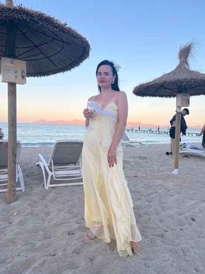 A woman stands on a sandy beach in Platja D'alcúdia, Spain, holding a glass of clear liquid. She is wearing a light yellow, flowing dress and has dark hair adorned with a white flower. She has red nail polish on her fingernails. The background shows a calm sea with a distant coastline and a wooden pier. Two thatched umbrellas are visible, one behind the woman and one to her right. Beach chairs are scattered on the sand. Several people are present in the background, some near the pier and others further away. The sky is a gradient of blue and pink, indicating it is either sunrise or sunset.