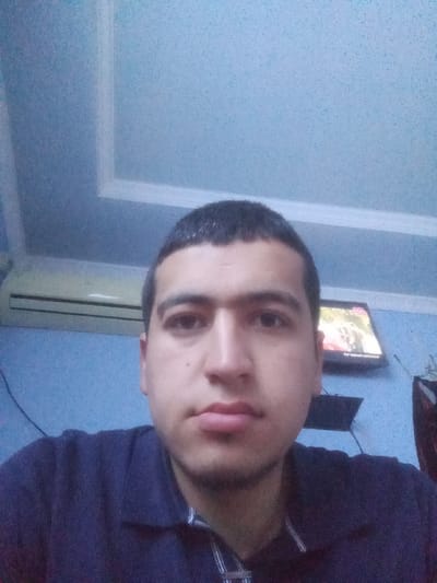 The image displays a head-and-shoulder shot of an adult male subject in an indoor setting, identified as a room in Kasan, Uzbekistan. The subject, positioned centrally, has short, dark hair and light facial stubble, and is wearing a dark blue polo shirt with a striped placket. He looks directly at the camera. The background consists of light blue, textured walls and a light blue ceiling featuring a white decorative rectangular trim in the upper left quadrant. A white wall-mounted air conditioning unit with dark horizontal vents is visible on the wall to the left of the subject. On the right, a flat-screen television is mounted on the wall, displaying an active image with red/orange elements and text at its base. Dark electrical wires are visible running along the wall, particularly near the air conditioning unit and below the television. A dark shelf or stand is present below the television, and a partial view of another object is visible on the far right side of the frame.