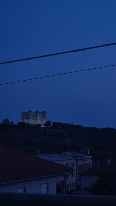 The image depicts a nighttime scene in Senj, Croatia, focusing on a castle on a hill. The sky is a deep, dark blue, indicating it is either dusk or dawn. Two power lines stretch across the upper portion of the frame, running horizontally. Below the sky, a dark, silhouetted hill rises, crowned by a stone castle. The castle appears to be a fortress with several prominent towers and crenellations. It is illuminated by artificial lights, making its structure visible against the dark sky. The base of the hill is covered in dark vegetation, with some scattered lights suggesting the presence of other structures or distant lights within the landscape. In the foreground, the roofs of houses are visible, also in silhouette, with some subtle details of their architecture hinting at a residential area. The overall impression is one of historical architecture set against a twilight sky.