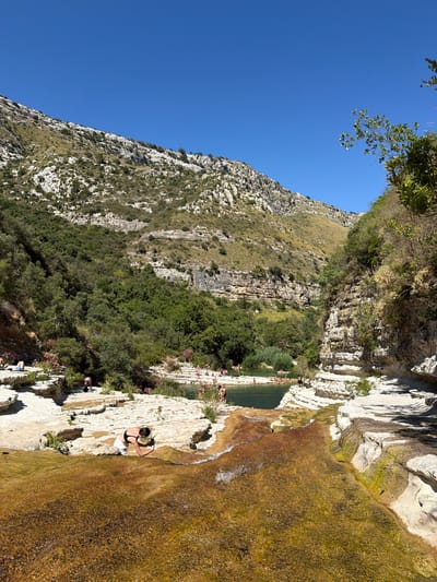 A clear, sunny day is depicted in a natural setting with a small waterfall or cascade flowing over rocky terrain into a tranquil pool. In the foreground, a woman in white shorts and a black bikini top kneels and appears to be taking a photo with her phone. Behind her, several people are visible in and around a green pool of water, some swimming and others relaxing on the rocky banks. Lush green trees and vegetation cover the slopes of the surrounding hills, which are characterized by layered rock formations and sparse greenery. The sky is a vibrant, cloudless blue. This natural area is identified as the Cavagrande del Cassibile, a nature reserve near Noto, Italy.