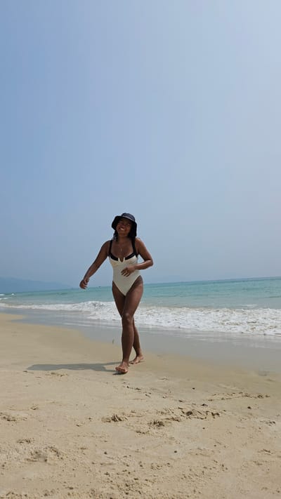 A full-body shot captures a smiling woman posing on a sunny beach in Bac Cam Ranh, Vietnam. The image depicts a vibrant outdoor scene with a clear, light blue sky on the left, meeting a turquoise ocean with white-foaming waves in the middle and right. Golden-brown sand stretches across the bottom right, wet near the water's edge.

The woman, with dark skin, stands barefoot on the wet sand. She is wearing a light-colored (possibly cream or off-white) one-piece swimsuit with dark shoulder straps and trim. A dark-colored bucket hat adorns her head. Her body is slightly angled, with her left arm extended towards the viewer and her right arm bent. She is looking directly at the camera with a wide, joyful smile, conveying a sense of happiness and relaxation. The sunlight highlights her skin and the details of the sand and water. No visible text is present in the image. The scene suggests a bright, pleasant day perfect for a beach outing.