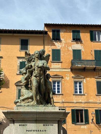 The image captures a vibrant street scene in Portoferraio, Italy, dominated by a large bronze monument set against a weathered building under a clear sky.

In the foreground, a substantial bronze sculpture depicts a complex grouping of numerous human figures, appearing intertwined or piled upon one another. The figures, rendered in a classical or heroic style, convey a sense of struggle or collective effort, some looking upwards, others in dynamic poses. The bronze has acquired a green patina characteristic of aged outdoor sculptures. The monument rests on a multi-tiered grey granite base.

Behind the monument stands a tall, light orange or ochre-colored building. Its facade shows signs of age with peeling paint and textured plaster. The building features multiple windows arranged irregularly across its surface. Many windows are equipped with dark green louvered shutters, some open, some closed, adding a characteristic Mediterranean touch. Some windows also have crisp white frames. A small, dark-colored balcony with intricate railings is visible on an upper floor, along with sections of decorative molding and exposed utility lines.

The bright sunlight from the clear sky casts strong shadows on the building's facade and the monument, suggesting a mid-morning or afternoon time. No particular activity is taking place; the scene is static, focusing on the architectural and sculptural elements.

Visible text on the granite base of the monument reads "MCMXV" (1915) and "MCMXVIII" (1918), indicating dates that likely refer to World War I. Below these dates, the word "PORTOFERRAIO" is clearly inscribed, confirming the location provided.