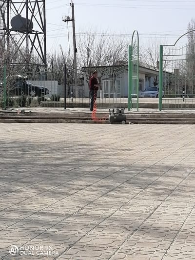 This image depicts an outdoor scene in the city of Samarkand, Uzbekistan, under clear skies with visible shadows. The foreground is paved with light-colored, rectangular interlocking pavers. A low concrete curb or step separates this pavement from a narrow concrete strip. On this strip, a small, gray portable air compressor with a coiled red hose is positioned. Behind the compressor, a light green metal fence, partially open with an arched gate, divides the scene. A male individual, dressed in a dark red long-sleeved shirt and dark pants, stands behind this fence, holding an object connected to the red hose. In the background, beyond the fence, a black sedan is partially visible to the left. To the right, a white building with windows and a white sedan parked in front of it are visible. The background also features numerous bare deciduous trees, utility poles with power lines, and a tall metal structure, possibly a water tower or cellular tower, with a dark tank at its apex on the far left.