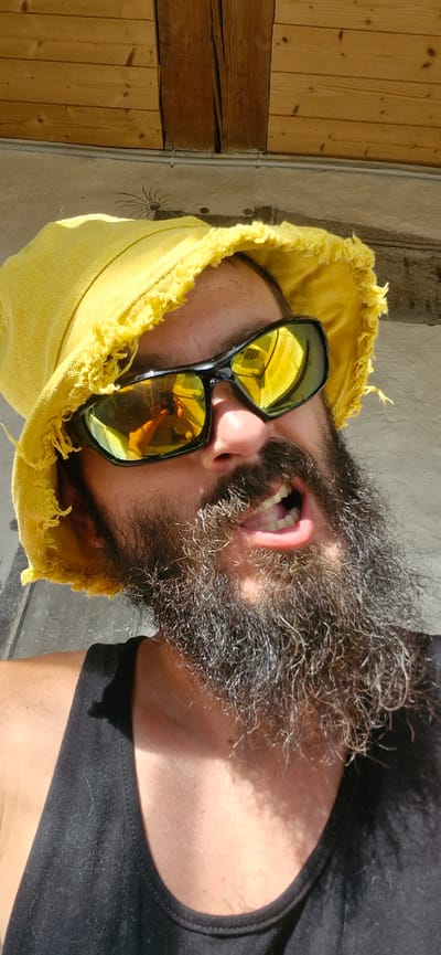 A man with a full beard and a yellow bucket hat is posing for a selfie outdoors. He is wearing sunglasses with yellow lenses that reflect the scene. His mouth is open, revealing his teeth. He is wearing a black tank top. The background consists of a wooden ceiling and a concrete wall with some graffiti. The image appears to be taken in Wattenwil, Switzerland, possibly at a rural or outdoor location given the building materials and natural light.