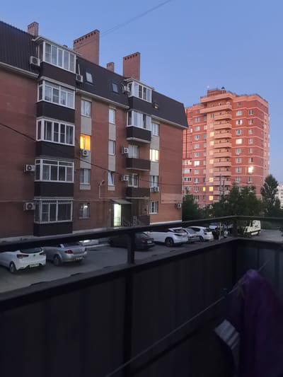 This media file shows an exterior view of apartment buildings in Verkhnetemernitsky, Russia, likely taken during dusk or early evening. The foreground features a balcony railing and some laundry hanging on a clothesline. Beyond the railing, a parking lot is visible with several cars, including a white Kia Rio, a silver sedan, and a white van. In the middle ground, a multi-story brick apartment building with numerous windows and balconies dominates the scene. One window on this building glows with warm light, suggesting indoor activity. Further in the background, a taller, modern apartment building with a distinctive peach-colored facade rises against the twilight sky. This building also has many illuminated windows. The sky is a clear, pale blue, indicating fair weather. The overall atmosphere is calm and residential. There is no discernible activity or event taking place beyond the cars parked and the general ambiance of a residential area at the end of the day. No people are clearly identifiable. License plates on the cars are partially visible but not legible enough to extract information.