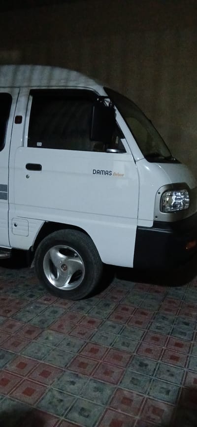 A white van with "DAMAS Deluxe" written on its side is parked on a tiled surface. The van is angled to the right, showing its front fender, headlight, and part of its windshield. The rear wheel, with a silver five-spoke rim, is visible. The background is dark and indistinct. The scene appears to be in a dimly lit outdoor area, possibly a street or driveway, in the city of Andijani, Uzbekistan.