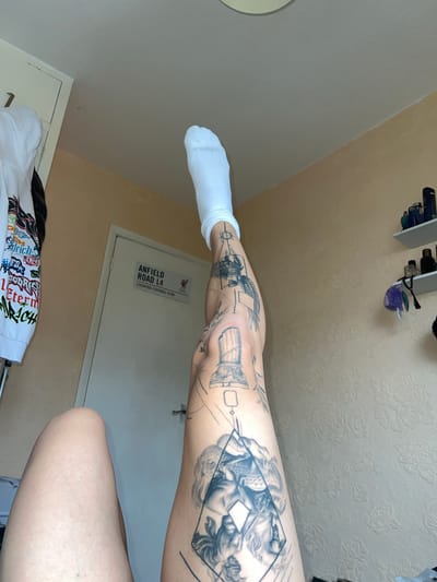 The image shows a person's leg with multiple tattoos. The leg is positioned in front of a white door with a sign reading "ANFIELD ROAD L4 LIVERPOOL FOOTBALL CLUB." The person is wearing a white sock. The location is not specified as Bristol, United Kingdom. The environment consists of a room with a patterned wall, a partially visible white garment, and some shelves with objects on them.