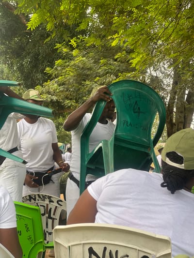 The image depicts an outdoor scene likely within Emure-Ekiti, Nigeria, characterized by lush green foliage overhead and in the background, suggesting a park, school grounds, or a similar open-air location. Several individuals, dressed in white t-shirts and green caps, appear to be participating in an organized activity.

In the foreground, a person is holding a green plastic chair, revealing graffiti written on its surface, including what appears to be "PLAISON" and other illegible markings. To the left, another green plastic chair is visible with markings that include "WAS HERE". In the mid-ground, a woman in a white t-shirt and green cap is seen wearing a black fanny pack around her waist. She is holding an orange device, possibly a phone or portable charger. Other individuals in white t-shirts and green caps are partially visible, some seated on white chairs. The overall atmosphere suggests a casual gathering or event in a semi-tropical environment.
