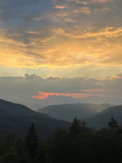 The image captures a dramatic mountain landscape at dusk in Katun Kobil do, Montenegro. The scene is dominated by a breathtaking sunset that paints the sky with vibrant hues. Thick, textured clouds are illuminated from behind by the setting sun, creating a golden glow that stretches across the left side of the frame. These bright clouds contrast with darker, more subdued cloud formations and the hazy, atmospheric layers of the distant mountains.

The right side of the image features a series of silhouetted mountain ranges that recede into the distance, creating a sense of depth. The slopes are densely covered with dark green coniferous trees, with some appearing as distinct shapes against the lighter sky. The foreground on the right is also filled with dark trees, their branches forming a textured silhouette.

The time of day is clearly sunset, indicated by the intense coloration of the clouds and the long shadows and atmospheric haze typical of this time. The weather appears clear enough for a dramatic sunset, though there's a noticeable atmospheric haze contributing to the layered effect of the mountains. The overall mood evoked is one of awe and tranquility, highlighting the natural beauty of the mountainous terrain at the close of day. No people, objects, or text are visible in the image.