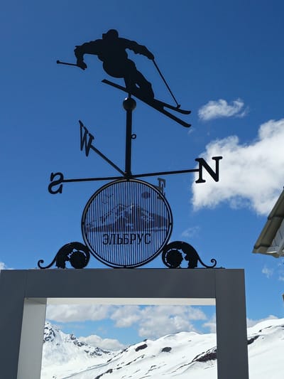 A black silhouette of a skier mid-jump is mounted on a weathervane against a bright blue sky with scattered white clouds. The skier's skis are angled downwards and to the right, and they hold ski poles. The weathervane also features directional indicators: "W" to the left, "N" to the right, and partially visible "S" to the bottom left. A circular ornament with vertical lines and the Cyrillic text "ЭЛЬБРУС" (Elbrus) is centrally located below the skier. Decorative black metal scrollwork is visible on either side of the circular ornament. The weathervane is mounted on a black pole. Below the weathervane, a portion of a modern, grey-framed structure is visible, framing a snow-covered mountain landscape. The mountains are white and rugged, with some exposed rock and patches of darker snow or ice. This scene is located in Terskol, Russia, likely at a viewpoint or resort area, given the prominent weathervane and the view of the snow-capped mountains.
