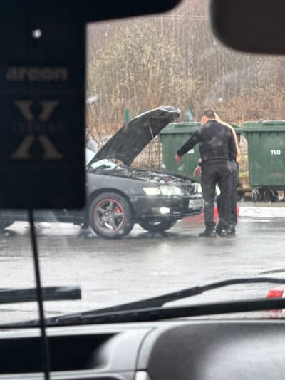 The image captures an outdoor scene, likely a street or parking area, under overcast daylight conditions, indicated by the wet ground. The primary subject is a man, dressed in dark clothing (black long-sleeved shirt and pants), standing beside a black car with its hood wide open. He appears to be leaning over, either inspecting or working on the car's engine.
In the background, two dark green dumpsters or waste bins are visible. One dumpster clearly displays the text "TKO" on its side. Beyond the bins, out-of-focus trees or brush create a natural backdrop.
The car itself is black, and one of its wheels, visible in the foreground, features a distinctive red accent on its rim. The overall setting suggests a car breakdown or maintenance in progress.
In the immediate foreground, a blurred object obstructs part of the view. Upon closer inspection, the text "Areeon XX" is visible on this object, which appears to be an air freshener, implying the photo was taken from inside another vehicle. The ground is consistently wet, suggesting recent rainfall. No strong emotional cues are evident from the man's posture, which indicates focus on the task.