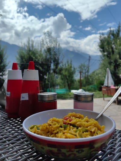 The image shows a close-up of a bowl of instant noodles with a fork resting on the side, placed on a woven metal table. In the background, two red squeeze bottles with white tops and labels reading "Red" are visible, along with a salt and pepper shaker. Further in the background, trees and mountains under a cloudy sky can be seen. The scene appears to be outdoors, possibly at a restaurant or a picnic spot. The presence of mountains suggests a location in a hilly or mountainous region. Chhyal, India is known for its scenic beauty and mountainous landscape, making it a plausible location for this scene.