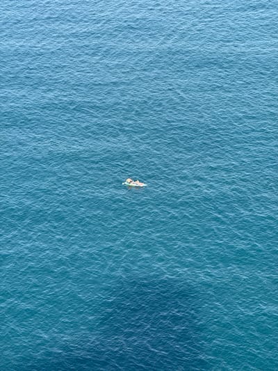 An aerial view captures a small, light-colored boat, likely a kayak or a small dinghy, floating in the center of a vast expanse of blue water. At least two individuals are visible within the boat, suggesting recreational activity such as kayaking or paddling.
The water's surface is covered in subtle ripples and small waves, reflecting the sunlight and creating a textured pattern across the entire frame. The dominant color of the water is a deep, rich blue with hints of turquoise, indicating clear coastal waters. The lighting suggests a bright, sunny daytime, with no visible shadows or cloud cover, implying calm and fair weather conditions.
The perspective is directly overhead, emphasizing the isolation of the boat and its occupants on the open water. There are no other boats, landmasses, or structures visible in the immediate vicinity, enhancing the sense of tranquility and remoteness. The scene is set in the coastal waters near Peníscola, Spain, as per the provided context. No text is visible in the image.