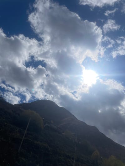 The image depicts a mountainous landscape in Juta, Georgia, under a partially cloudy sky with strong sunlight. The upper two-thirds of the frame are dominated by a bright blue sky interspersed with large, white and grey cumulus clouds. The sun is a prominent, intensely bright light source in the upper right quadrant, partially obscured by clouds, emitting a strong lens flare. The lower third of the image features a dark, steep mountain slope, largely in shadow, covered with sparse green and brown vegetation. Visible on the mountain are several thin, vertical poles, likely utility or fence posts, particularly on the left. Approximately mid-slope, towards the center-right, a multi-story building with a flat roof and multiple windows is discernible. No people are visible.