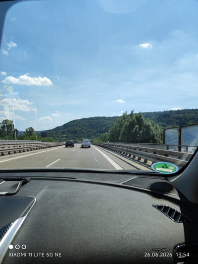 The image shows a view from inside a car driving on a highway in Unterurbach, Germany. The road is multi-lane and separated by concrete barriers. There are other vehicles on the road, including a dark-colored car and a white van with "toom8" written on its side. The landscape outside the car is lush and green, with rolling hills covered in trees. The sky is blue with scattered white clouds, indicating a clear and pleasant day. The time of day is indicated as 13:54 on June 26, 2026, suggesting it is mid-afternoon. A circular sticker with German text and a number "4" is visible on the windshield. The dashboard of the car is in the foreground, with a carbon fiber trim and the text "XIAOMI 11 LITE 5G NE" and the date and time stamped on the bottom left and right of the image. The overall mood of the image is one of travel and leisure, capturing a moment on a sunny day during a road trip.