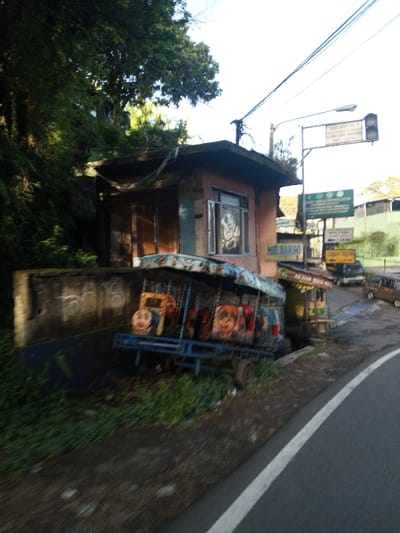 The scene depicts a roadside area, identified as being in the city of Khouathineung, Laos. Visible signage, however, contains text in Indonesian/Sundanese, specifically "MIE BAKSO," "MIE AYAM BAKSO MAS," "JAGA JARAK 150 M" (Keep Distance 150M), and "LEMBAH TAQWA 1.4 KM."
On the left, a small, two-story structure with a faded pinkish-orange facade and a flat, dark-colored concrete roof is partially concealed by dense green foliage from an overgrown embankment. The building features a single window with a metal grate and an internal graphic design. Adjacent to this structure, a blue-painted wheeled trailer-like object, likely a stationary or dismantled children's ride, is positioned. It has a light-colored, patterned tarp canopy and multiple child-sized seats painted with cartoon characters, including one resembling Thomas the Tank Engine.
To the right of the trailer, a small wooden food stall with a brown corrugated roof is visible, displaying signs for "MIE BAKSO" and "MIE AYAM BAKSO MAS." Above and behind these foreground objects, a metal pole supports a functioning traffic light, illuminated red, and multiple directional signs. These signs include "JAGA JARAK 150 M," another indicating "LEMBAH TAQWA 1.4 KM," and a partially visible green sign. Overhead power lines are stretched across the sky.
The right side of the image features a paved asphalt road with a solid white line marking its edge, curving gently into the distance. Two dark-colored vehicles are discernible further down the road, and an older, lighter-colored vehicle is visible beyond them to the right. The immediate foreground and areas around the structures consist of bare earth and sparse green vegetation. No individuals are present in the image. The lighting suggests daylight conditions.