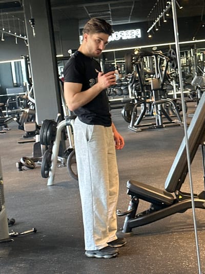 An adult male stands in a gym located in Jestetten, Germany. The individual, with dark, styled hair and facial stubble, wears a black short-sleeved t-shirt, light grey sweatpants, and dark athletic shoes with segmented soles. He holds a black smartphone in his raised right hand, consistent with the act of capturing a self-portrait in a mirror. His gaze is directed towards the camera's perspective. The gym environment features dark rubberized flooring and dark walls. In the background, reflected in a large mirror, are various pieces of exercise equipment including weight racks, cable machines, and benches. A reversed, illuminated sign displaying "PERFORMA" is visible on the distant wall's reflection. A black adjustable weight bench is partially present in the lower right foreground.