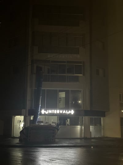 The image depicts a multi-story building at night in Engomi Lefkoşa, Cyprus. The scene is dark, with artificial light emanating from within the building and a prominent outdoor sign.

The building features several windows, some of which appear to have blinds or shutters. On the right side of the building, a large, vertical, illuminated sign is clearly visible. The sign displays the name "INTERVALA" in bright white text, preceded by a cross or plus symbol. Below this, the text "Rehab & Fitness Centre" is written, indicating the nature of the business.

The environment appears wet, with a noticeable sheen on the dark wall to the far right, suggesting recent rain or damp conditions. Some dark pipes or conduits run diagonally across the lower central portion of the image. No people are visible, and no specific activity is taking place other than the static presence of the building and its illuminated sign. The overall atmosphere is quiet, typical of an urban setting after dark.