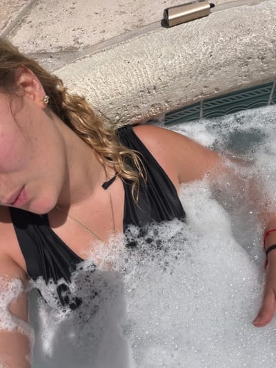 A woman is reclining in a hot tub filled with bubbles. The hot tub is located on a patio with a stone wall and green tile detail. The woman is wearing a black bikini top and a gold necklace. A vape pen is resting on the edge of the hot tub. The location is presumably in Key Biscayne, United States of America.