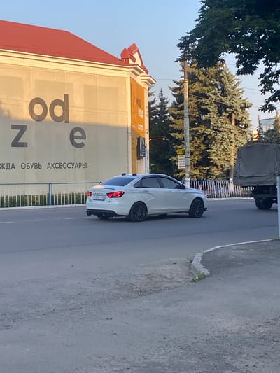 The image captures a street scene in the city of Antratsyt, Ukraine, bathed in the warm glow of late afternoon sunlight. On the left, a building with a prominent red roof displays a large sign with the letters "odze" in dark gray, followed by text in a lighter shade indicating "ЖДА ОБУВЬ АКСЕССУАРЫ" (clothing, shoes, accessories).

A white Lada Vesta sedan, bearing the license plate number "6529 TT 65", is driving on the asphalt road in the foreground. The car has tinted windows and black rims. To the right of the sedan, a grey canvas-covered truck is parked or moving slowly on the same road.

The environment is urban, with a clear blue sky above. Tall green evergreen trees are visible behind the building and along the street, interspersed with utility poles and wires. A white metal fence runs along the edge of the sidewalk. The foreground shows a paved road with a concrete curb and some patches of dry grass and small stones. The overall impression is of a quiet street on a sunny day.