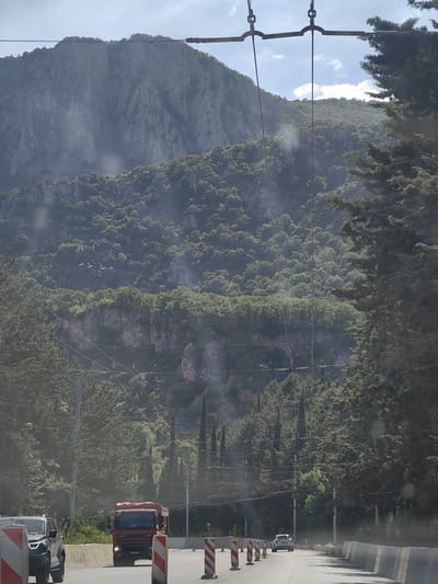 The image depicts a sunlit scene with a road winding through a mountainous, tree-covered landscape. In the foreground, a red truck with a "DAF" logo is visible on the road, its license plate reading "B 678 KA 90". Next to it, a dark SUV is also on the road, partially obscured by construction barriers. The road itself appears to be undergoing some form of construction or repair, indicated by the presence of white and red striped barriers and concrete barriers lining the side.

Behind the road, a dense forest covers the lower slopes of a massive, rugged mountain. The mountain's rocky face is prominent, with visible crevices and sharp angles. Overhead, power lines and what appear to be trolleybus wires crisscross the sky, suggesting an area with public transportation infrastructure. Tall, dark green coniferous trees line the right side of the road, partially obscuring the view of the mountain and sky.

The time of day appears to be daytime, with bright sunlight casting shadows and creating lens flares in the image. The weather seems clear, with some scattered clouds visible in the bright blue sky. The overall impression is of a scenic route through a natural landscape, possibly near a mountainous region or a significant elevation change, with ongoing infrastructure work impacting the immediate road area. No people are clearly visible. The location is identified as Kiparisnoe, Ukraine.