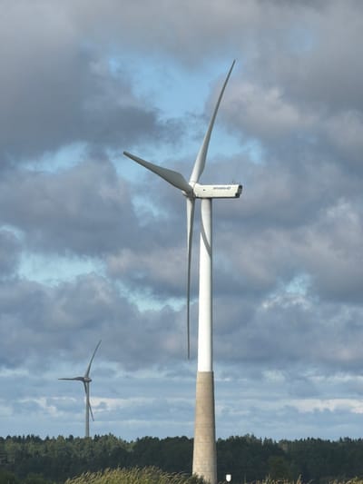 The image depicts a landscape scene with two wind turbines situated in Koogu, Estonia. In the foreground, a tall white wind turbine with a concrete base stands prominently, its blades angled upwards. To the left and further in the background, a second, smaller wind turbine is visible, also with its blades in an upward position. The turbines are set against a backdrop of a dense green forest line. Above the forest, the sky is filled with a mix of dark gray and light blue clouds, indicating an overcast or partly cloudy day. The ground in the immediate foreground is covered with dry grass.