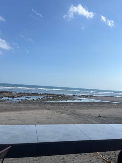 This is a photograph of a beach in Pekutatan, Indonesia. The sky is a clear blue with a few scattered white clouds. The ocean is a deep blue, with gentle waves breaking on the shore. The beach is covered in dark sand, with a scattering of rocks and driftwood. In the foreground, there is a tiled ledge, suggesting this is a vantage point overlooking the beach, perhaps a cafe or seating area. The tide is out, revealing shallow pools of water among the rocks. The overall impression is one of a serene and natural coastal landscape.