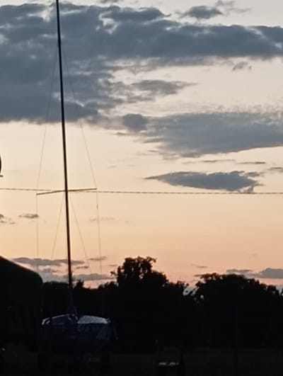 The image depicts a sailboat mast and rigging silhouetted against a dusky sky filled with clouds. A horizontal power line stretches across the frame. In the foreground, a dark silhouette of trees and foliage is visible, with a portion of a dark structure on the left. A small sailboat, also in silhouette, rests on land in the lower center of the image. The scene appears to be set in a location near Sztynort, Poland, likely at dusk or dawn, given the lighting conditions and the colors of the sky. No people are visible.