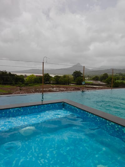 The image depicts an outdoor infinity pool overlooking a lush, green landscape in Ajivali, India. The pool's edge is visible in the foreground, with bright blue tiles lining its sides. The water in the pool is clear and reflects the overcast sky above. Beyond the pool, a wire fence stretches across the frame, separating it from the natural environment.
The landscape features a body of water in the middle ground, likely a river or lake, with trees and vegetation lining its banks. Further in the distance, rolling hills and mountains can be seen under a cloudy sky. The ground between the pool and the water appears to be muddy and disturbed, possibly due to recent rain. Two wooden poles with wires attached are also visible, adding to the rustic outdoor feel of the scene. The overall atmosphere is serene and natural, with the modern infinity pool providing a luxurious contrast to the surrounding environment.