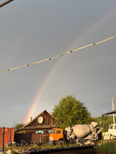 This is a landscape photograph taken in Skovorodino, Russia. The scene depicts a rural yard with a wooden house, a cement mixer truck, and other miscellaneous objects. A prominent rainbow arches across the overcast sky. The house is a single-story structure with a wooden exterior and a satellite dish mounted on the roof. An orange cement mixer truck is parked in front of the house. The yard is cluttered with various items, including what appear to be motorcycles or scooters, scrap metal, and possibly a shipping container. A utility pole with wires is visible in the background. The overall atmosphere suggests a post-rain or stormy weather condition, indicated by the rainbow and the subdued lighting.