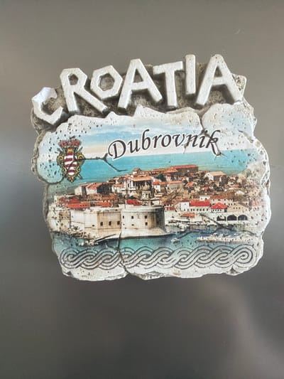 A magnet featuring the word "CROATIA" in raised white letters on a textured, off-white background is displayed. Below this, a colorful image depicts the city of Dubrovnik, Croatia, with its historic stone buildings, red-tiled roofs, and a body of blue water. A coat of arms with red and white stripes and a crown is visible on the left side of the image. The bottom of the magnet has a decorative border resembling intertwined waves. The magnet is positioned on a dark, flat surface, likely a refrigerator. There are no people or animals present in the image. The magnet is also not located in Sderot, Israel.