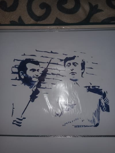 The image displays a blue ink drawing on a white surface, likely paper or a poster. The drawing features two men, rendered in a sketch-like style with dark blue lines. The man on the left is depicted from the chest up, with a beard and a neutral expression. He is holding a thin, rod-like object. The man on the right is shown in a similar pose, with his face turned slightly away from the viewer, and his hand is visible holding a cigarette or a similar object. The background of the drawing is abstract, with horizontal and vertical lines suggesting a brick wall. The overall presentation is simple and lacks any specific urban or environmental context that would indicate a location in Gulbag, Uzbekistan. The image is captured in a vertical orientation, and the top portion of the image shows a textured surface, possibly a rug or upholstery.