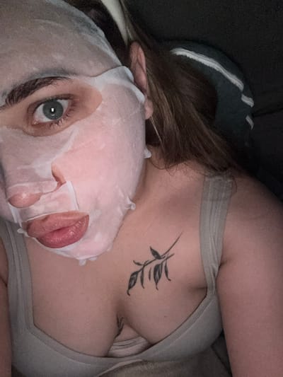 A close-up, overhead shot shows a woman with a sheet mask on her face and a tattoo on her chest. She is wearing a light gray tank top. The sheet mask covers most of her face, leaving her eye, nose, and mouth visible. Her eye is green and her lips are pink. The tattoo on her chest is a black, minimalist design of a branch with leaves. Her hair is brown and pulled back. The background is dark and out of focus, suggesting an indoor setting. The scene does not provide any specific information or context that would allow for its location in Breisach am Rhein, Germany.