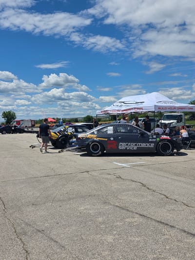The image depicts a sunny day at a race track, likely in or near Akatsievo, Bulgaria, given the context. The foreground shows a dark grey, highly modified race car with "PERFORMANCE SERVICE" prominently displayed on its side. The car has large racing slicks with "Hoosier" branding and a roll cage visible through the windows. Another race car, with yellow and black graphics and the number "802", is positioned behind and to the left of the primary car. A white tent, branded with "VASTAL TEAM CAR" and "BUILD YOUR DREAM CAR", is set up behind the cars, providing shade for a group of people. Several individuals are gathered under and around the tent, some appearing to be spectators or crew members. A white van is parked behind the tent. In the background, a red fire truck is visible, along with other vehicles and a grassy area with some trees. The sky is a vibrant blue with scattered white clouds. The ground is an asphalt surface typical of a race track or parking lot. A person in a dark t-shirt and grey shorts is walking away from the camera towards the race cars.