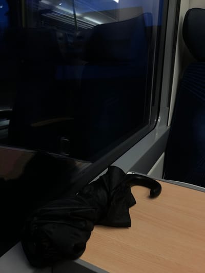 An umbrella rests on a light brown surface next to a train window. The window reflects dark shapes, possibly from the opposite seat. A blue and black striped seat is visible in the upper left corner. The umbrella is black and appears folded or crumpled. The window frame is gray. The overall perspective is slightly tilted.