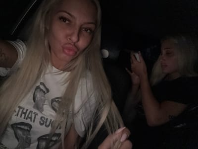 Two women are present in the image. One woman, positioned closer to the camera, has long blonde hair and is wearing a white t-shirt with a graphic print. The other woman, further away, is wearing a black top. The environment appears to be indoors. There is no indication of the location in Cherna Gora, Bulgaria.