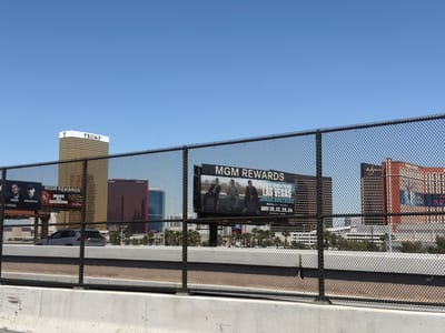 The image depicts a scene in Paradise, Nevada, USA, where various hotel and advertising signs are visible behind a chain-link fence and concrete barrier. The foreground is dominated by the fence and barrier, while in the background, several prominent buildings are featured. These include the Trump International Hotel Las Vegas, the Wynn, and Encore hotels, along with advertising billboards for MGM Rewards featuring DJs and events. The billboards display promotional content, mentioning names like Tiesto and Dom Dolla, and dates for events in Las Vegas. The sky is clear and blue. A white or light-colored car can be seen driving behind the fence.