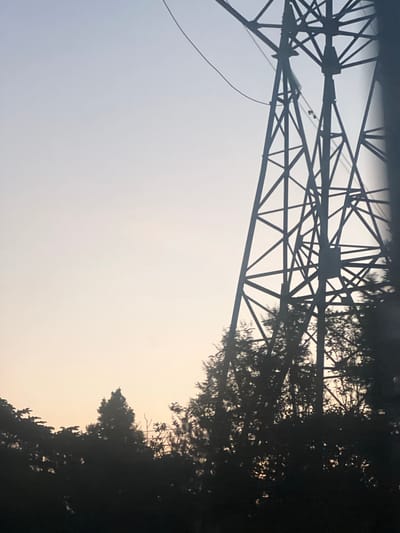 The image depicts a silhouette of a power transmission tower against a pale sky, with the dark silhouettes of trees on the right side. The sky transitions from a light blue or gray at the top left to a pale yellow or orange hue towards the bottom right, suggesting either sunrise or sunset. The power tower is a complex metal structure made of intersecting beams and supports, casting a sharp, angular shadow. A thin wire hangs from the tower, disappearing into the dark foliage. The trees are densely packed and their branches create a feathery, organic contrast to the geometric lines of the tower. The overall impression is one of industrial infrastructure juxtaposed with nature, possibly taken in a rural or semi-rural setting. The lighting suggests it is either early morning or late evening. There is no visible text in the image. The location context of Bol'shoe Sareevo, Russia, places this scene in a specific geographic area.