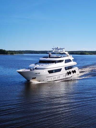 A large, modern multi-deck motor yacht, predominantly light grey and white, navigates calm, deep blue water in Novodevich'e, Russia. The vessel is positioned centrally, moving from right to left, generating a distinct white bow wave at its front and a frothy wake behind its stern. Several black cylindrical fenders are visible along the starboard side of the yacht. The yacht features multiple large rectangular windows on its main and upper decks, and an array of communication and navigation equipment is mounted on the uppermost deck. In the distant background, a dense, verdant forest lines the shores on both sides of the wide waterway. The sky above is clear and bright blue, with a few faint white contrails present. The scene is brightly illuminated by direct sunlight. No individuals are clearly identifiable aboard the yacht or in the surrounding environment.
