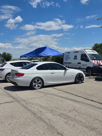 The image captures an outdoor scene, likely at a car event or service area, in the city of Akatsievo, Bulgaria. In the foreground, a white BMW coupe is parked on an asphalt surface. The car has custom black and silver rims and "Hoosier" branding on its tires. To the left, a white hatchback, possibly a Seat Leon, is partially visible.

Behind the cars, a large blue canopy tent is set up, suggesting an event or covered area. Further in the background, a white van with "TRUCK SERVICE" branding is parked. The van displays text listing services like "VOLVO RENAULT & TRAILER REPAIR," "ECU SOFTWARE UPDATE," "ROADSIDE ASSISTANCE," and "DIAGNOSTICS." Contact phone numbers are also visible on the van.

The environment is characterized by a bright, sunny day with a blue sky filled with scattered white clouds. Trees and foliage are visible in the background, indicating a semi-rural or suburban setting. Tire tracks are visible on the asphalt in the foreground, hinting at car activity. No people are clearly visible in the frame.